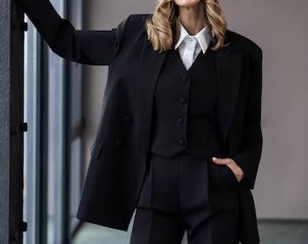 May include: A black three-piece suit consisting of a structured blazer, a button-up vest, and tailored trousers. The ensemble is paired with a crisp white collared shirt featuring a single visible button at the neck, creating a formal appearance.