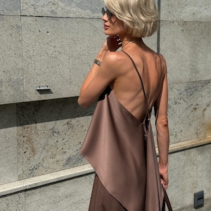 以下が含まれることがあります： ブラウンのサテンのドレス、細いストラップとドレープされた非対称のデザイン。 金髪とサングラスをかけた人が、テクスチャ加工された灰色の壁の前に立ってドレスを着ています。 手首には時計が見えます。