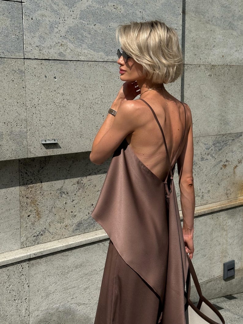 以下が含まれることがあります： ブラウンのサテンのドレス、細いストラップとドレープされた非対称のデザイン。 金髪とサングラスをかけた人が、テクスチャ加工された灰色の壁の前に立ってドレスを着ています。 手首には時計が見えます。