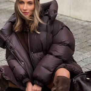 Puede incluir: Una chaqueta acolchada marrón con una gran capucha y un sombrero de pana marrón a juego. La chaqueta tiene un diseño acolchado y cierre de cremallera. La persona también lleva botas de ante marrones y un bolso de cuero marrón.