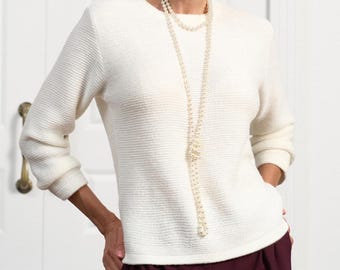May include: A long-sleeved, off-white sweater with a horizontal ribbed texture and a round neckline, paired with a long, multi-strand pearl necklace featuring a central knot detail and dark maroon trousers.