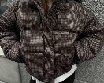 May include: Brown and black houndstooth puffer jacket with a hood and a zipper closure. The jacket has two front pockets and a quilted design. The model is wearing a white turtleneck and black leggings.