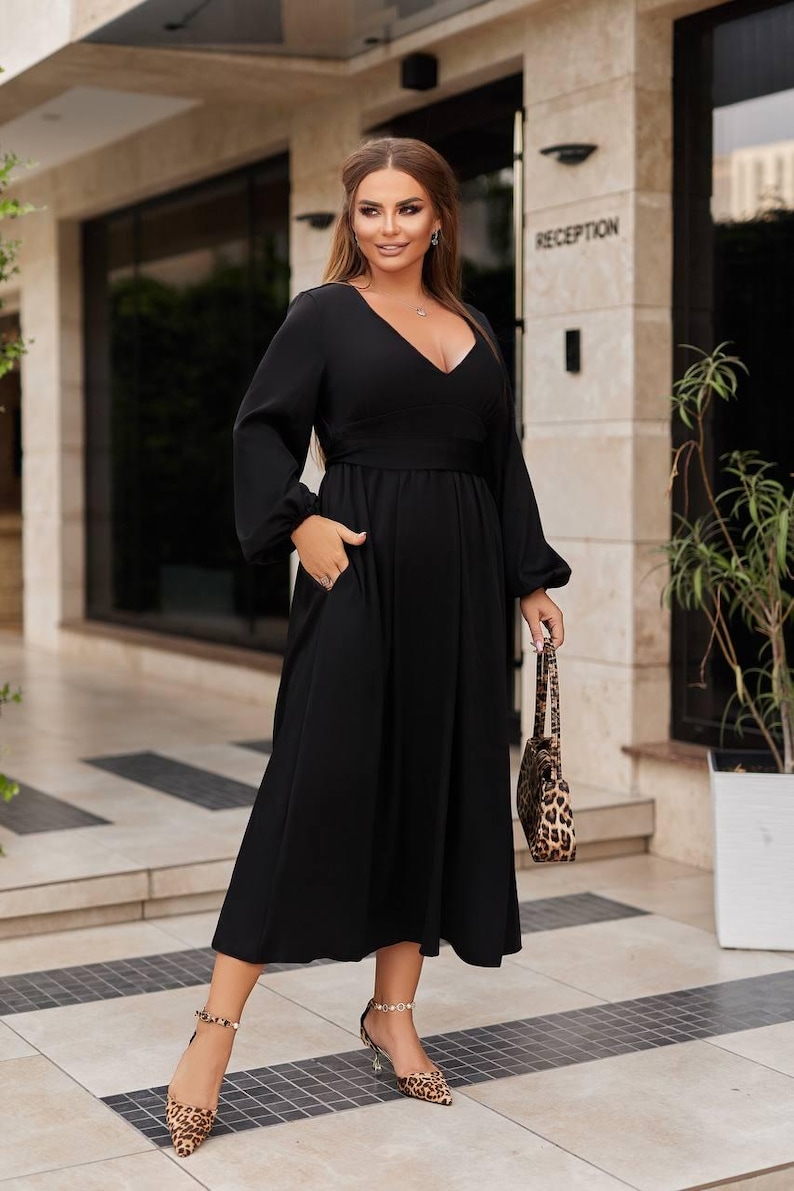 May include: A black midi dress with long sleeves and a V-neck. The dress has a fitted waist and pockets. The person is carrying a leopard print handbag and wearing leopard print heels. The word "RECEPTION" is visible in the background.