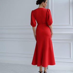 May include: A striking red dress with a fitted bodice and a flared skirt. The dress has short, puffed sleeves and a V-neck. The hemline is below the knees. The model is wearing black high-heeled sandals.