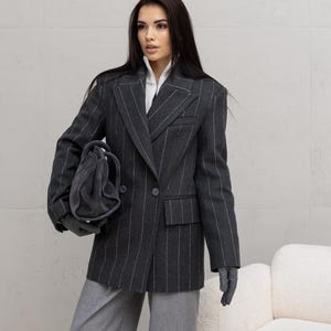 Pode incluir: Um blazer cinza escuro com riscas finas e design de abotoamento duplo, combinado com calças cinza a condizer. O blazer apresenta riscas finas brancas e um fecho de dois botões. Uma mala e luvas cinzentas completam o conjunto. Uma versão moderna da alfaiataria clássica.