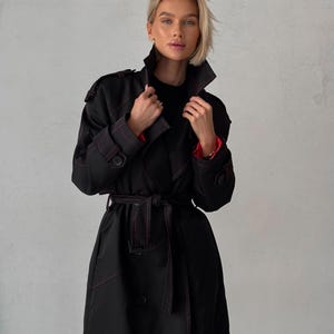 May include: A black trench coat with red stitching. The coat has a double-breasted closure, a belt, and a collar.