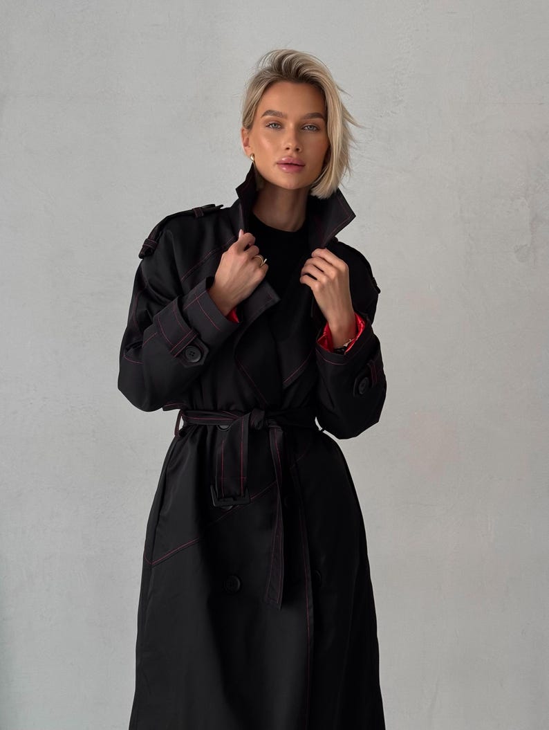 May include: A black trench coat with red stitching. The coat has a double-breasted closure, a belt, and a collar.