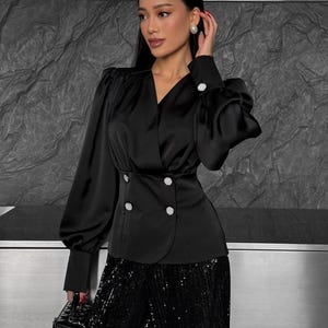 May include: A black satin blouse with a v-neckline, long sleeves, and a double-breasted button closure. The blouse has a fitted waist and a slightly flared hem. The model is wearing black sequined pants.