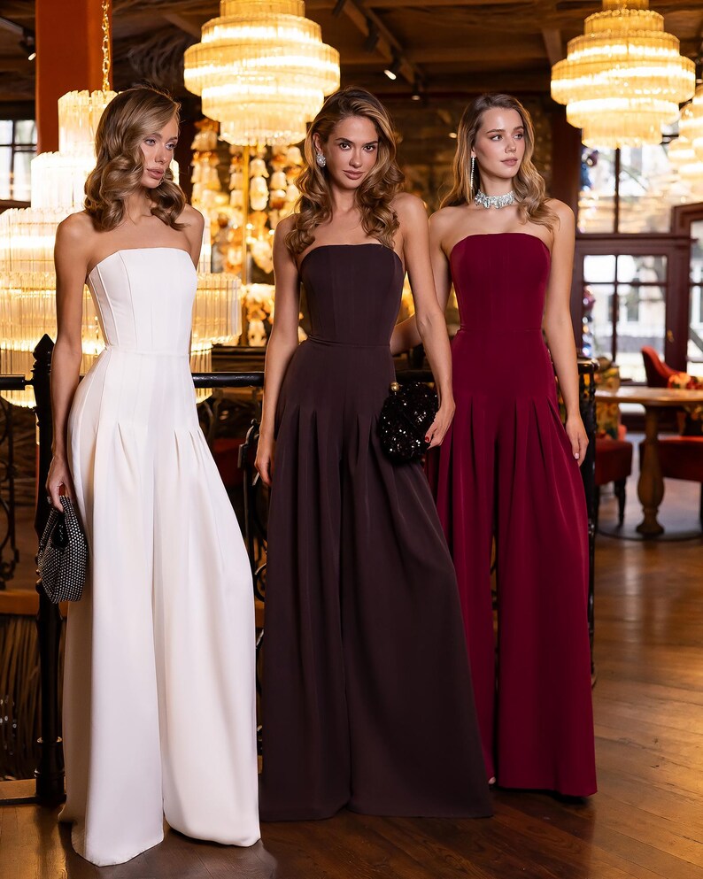 May include: Three strapless jumpsuits featuring fitted bodices and wide-leg trousers. One jumpsuit is white, one is dark brown, and one is deep burgundy. Each garment has a clean, minimalist silhouette with a straight neckline and floor-length hem.