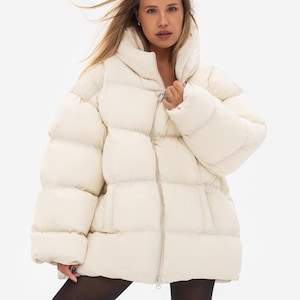 May include: An oversized cream-coloured puffer jacket featuring a high collar, horizontal quilted panels, and a full-length front zip closure. The jacket has long, wide sleeves and a boxy silhouette, constructed with a smooth, matte-finish fabric.