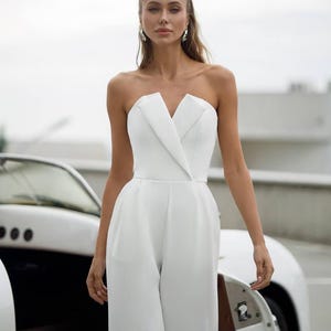 May include: A strapless white jumpsuit featuring a structured bodice with a notched lapel detail at the center of the neckline. The garment has a fitted waistline that transitions into wide-leg trousers, creating a clean and tailored silhouette.