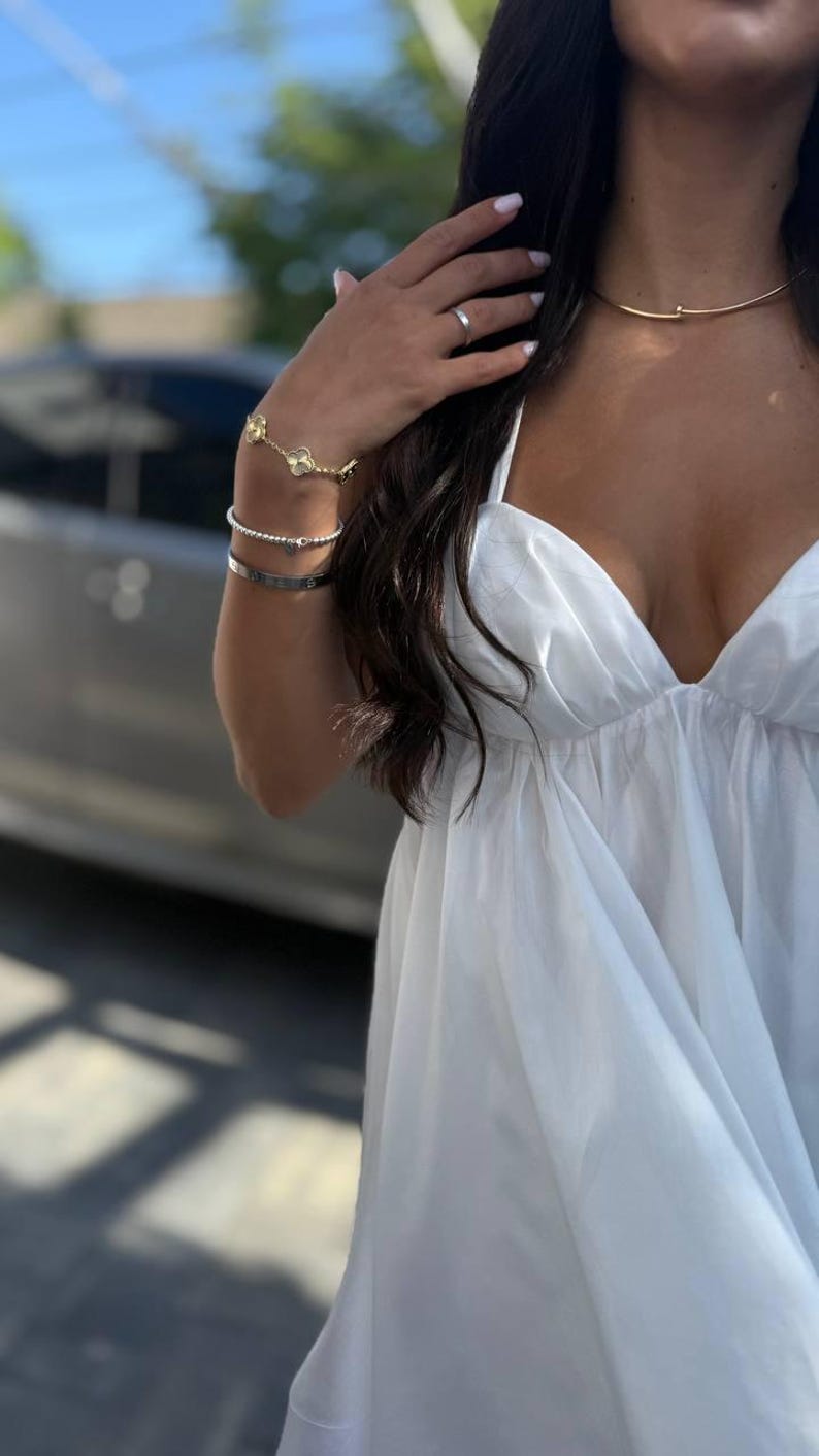 Puede incluir: Un vestido blanco con escote pronunciado se combina con joyas doradas, incluyendo un collar y una pulsera con dise&ntilde;o de tr&eacute;bol. Una pulsera plateada y un anillo completan el look. La imagen est&aacute; tomada al aire libre.