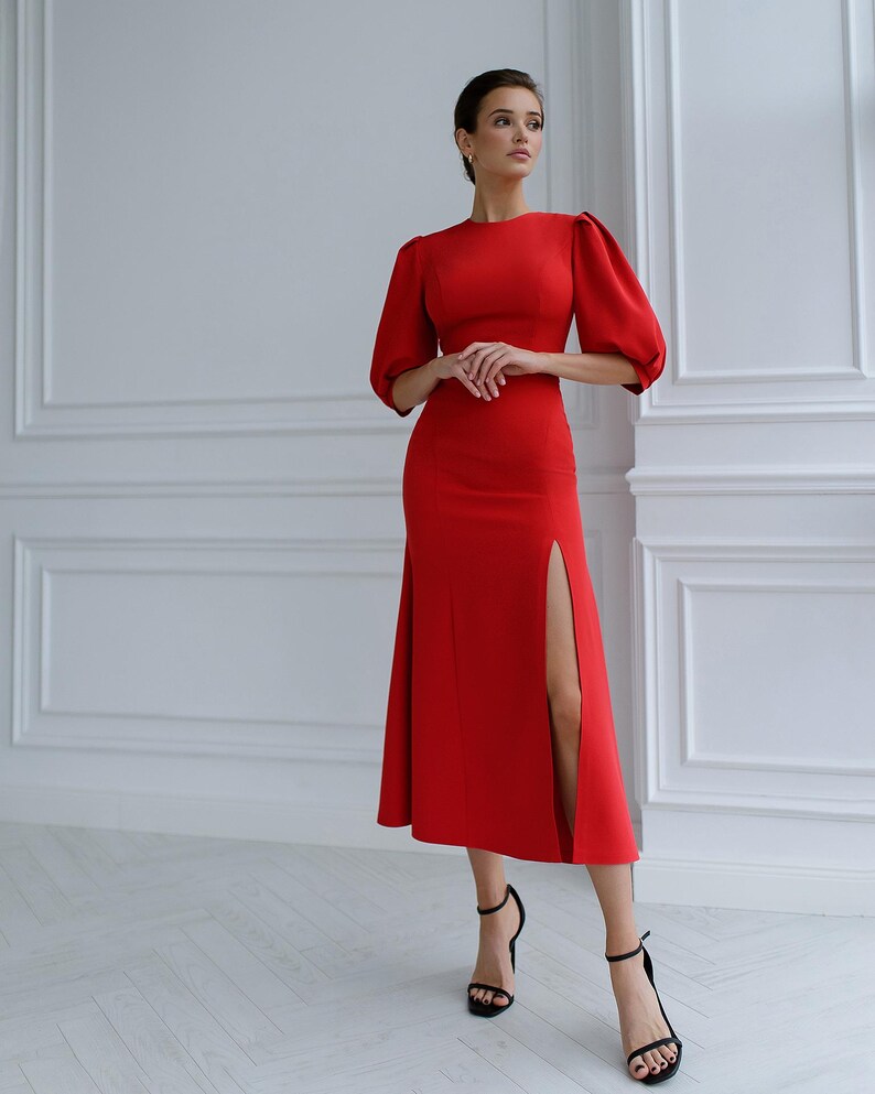 May include: A striking red dress with a knee-length skirt and a high slit. The dress has short, puffed sleeves and a round neckline. The model is wearing black strappy heels. The background is a white wall.