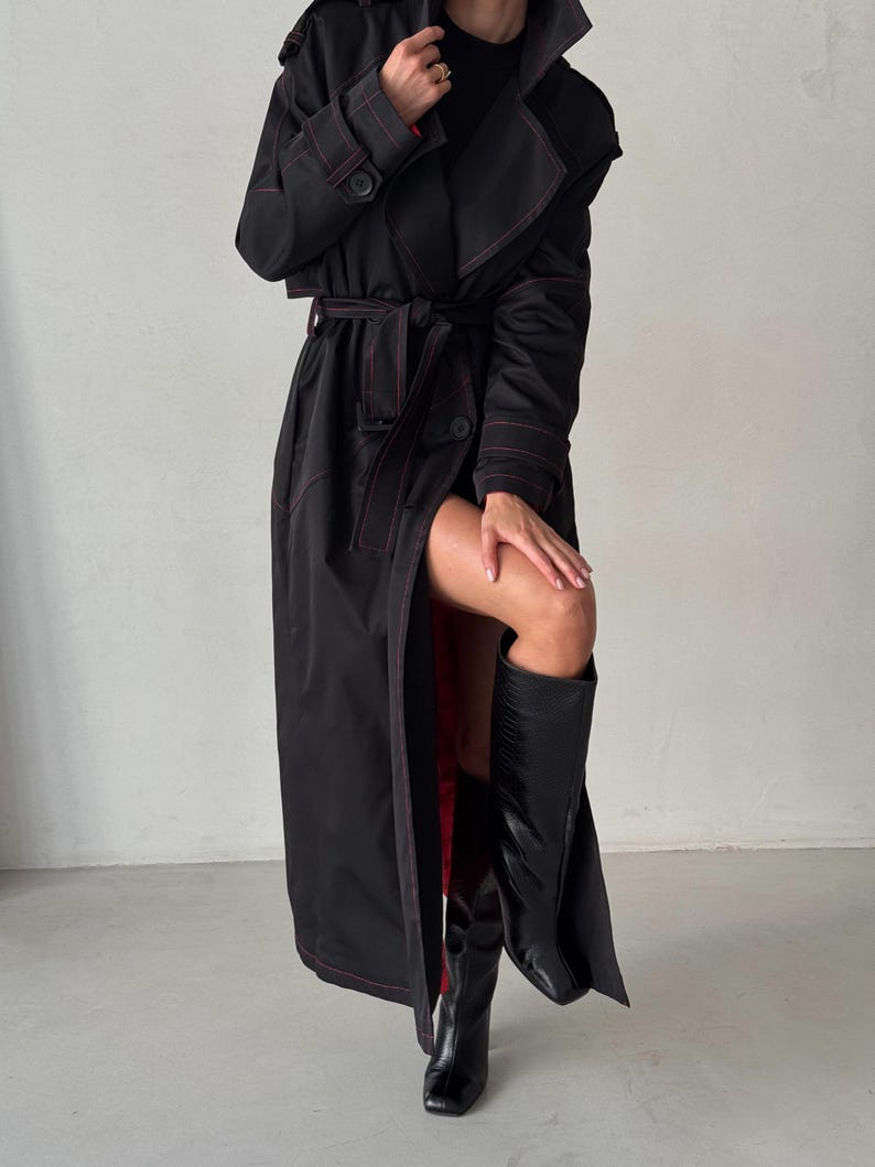 May include: A long black trench coat with red stitching. The coat has a belt and a large collar. The model is wearing black boots.