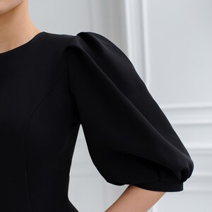 May include: A black dress with a round neckline and elbow-length, puffed sleeves. The dress has a fitted bodice and a simple design, suitable for formal occasions. The image shows the sleeve detail.