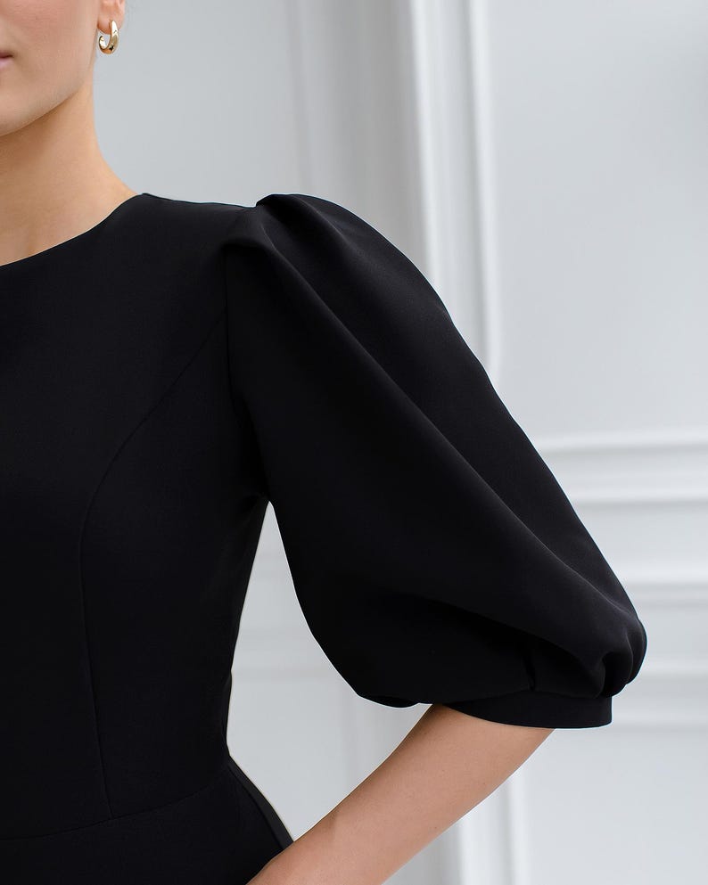 May include: A black dress with a round neckline and elbow-length, puffed sleeves. The dress has a fitted bodice and a simple design, suitable for formal occasions. The image shows the sleeve detail.
