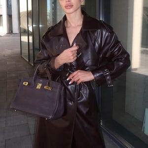 Puede incluir: Una mujer con un abrigo de cuero marr&oacute;n con cintur&oacute;n y un bolso de mano de cuero marr&oacute;n. Lleva gafas de sol y tiene el pelo recogido hacia atr&aacute;s.