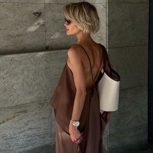 以下が含まれることがあります： 茶色のサテンのトップスと、お揃いのワイドパンツを着た女性。サングラス、時計、ネックレスも着用しています。茶色のストラップが付いた白いトートバッグが肩にかけられています。背景は、テクスチャ加工された灰色の壁です。