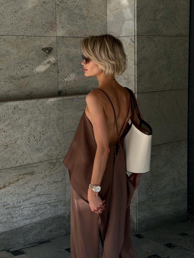 以下が含まれることがあります： 茶色のサテンのトップスと、お揃いのワイドパンツを着た女性。サングラス、時計、ネックレスも着用しています。茶色のストラップが付いた白いトートバッグが肩にかけられています。背景は、テクスチャ加工された灰色の壁です。