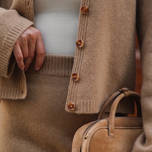 May include: A tan knit cardigan with brown marbled buttons worn over a cream-colored top, paired with a matching tan knit skirt and a small, structured tan suede handbag with a top handle and a zipper closure along the top edge.