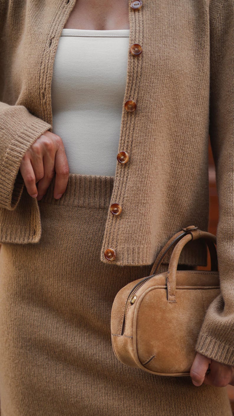 May include: A tan knit cardigan with brown marbled buttons worn over a cream-colored top, paired with a matching tan knit skirt and a small, structured tan suede handbag with a top handle and a zipper closure along the top edge.