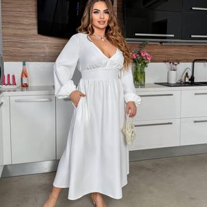 May include: A white, long-sleeved dress with a V-neck and a fitted waist. The dress features a flowing skirt and pockets. The person is wearing silver pointed-toe heels and holding a small, beaded purse. The background is a modern kitchen.