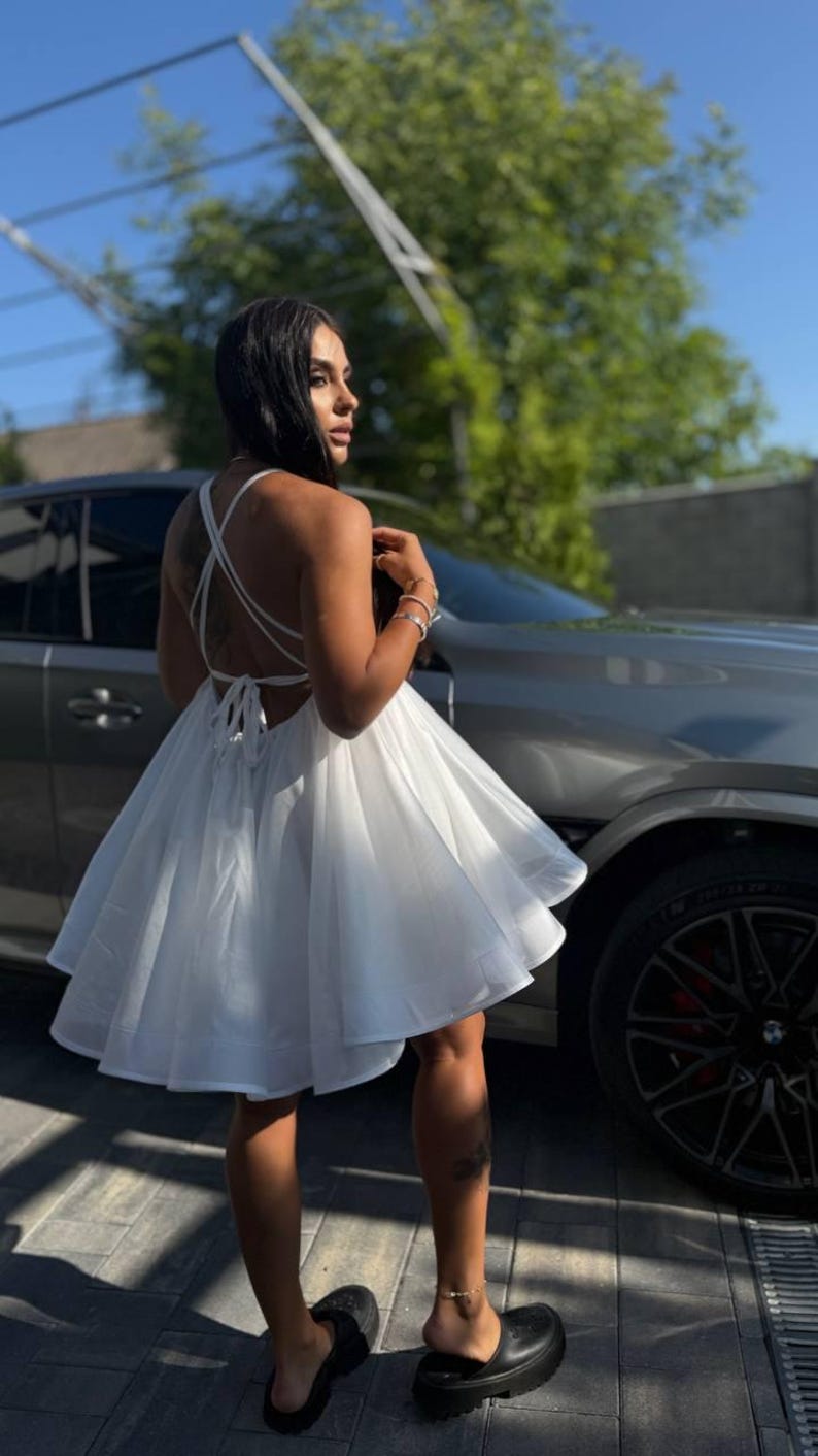 Puede incluir: Un vestido blanco sin mangas con falda acampanada y detalle de espalda cruzada. El vestido se lleva con sandalias de plataforma negras. La imagen est&aacute; tomada al aire libre, con un coche al fondo.