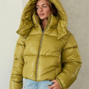 May include: A chartreuse puffer jacket with a high collar and an oversized, detachable hood. The jacket features a front zip closure, horizontal quilted stitching, and long sleeves with elasticated cuffs, paired with light blue denim jeans.