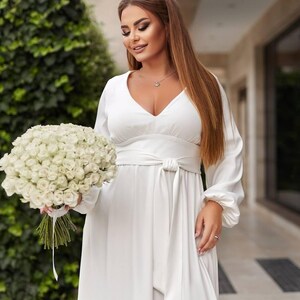 May include: A white, long-sleeved dress with a V-neck and a tie-waist. The dress is paired with a bouquet of white roses. The overall theme is bridal or formal wear.