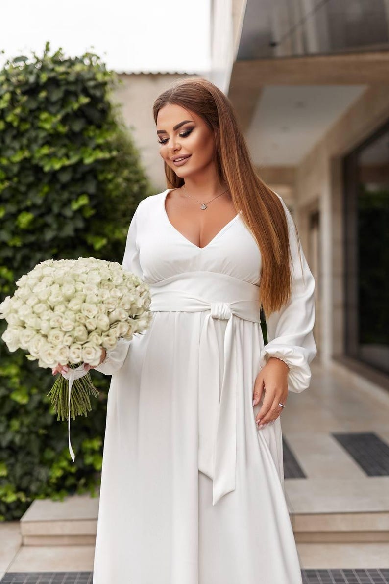 May include: A white, long-sleeved dress with a V-neck and a tie-waist. The dress is paired with a bouquet of white roses. The overall theme is bridal or formal wear.