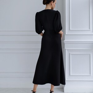 May include: A black dress featuring a V-neck and elbow-length sleeves. The dress has a fitted bodice and a flowing skirt. The model is wearing black high-heeled sandals. The background is a white wall with decorative panelling.
