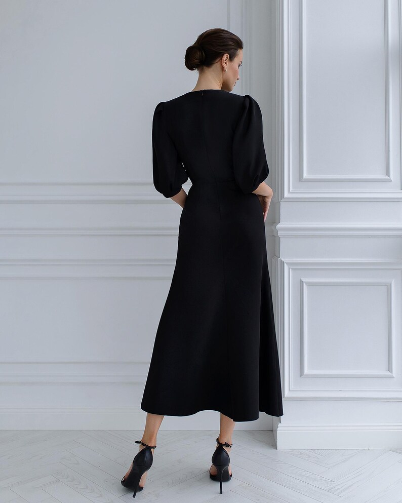 May include: A black dress featuring a V-neck and elbow-length sleeves. The dress has a fitted bodice and a flowing skirt. The model is wearing black high-heeled sandals. The background is a white wall with decorative panelling.