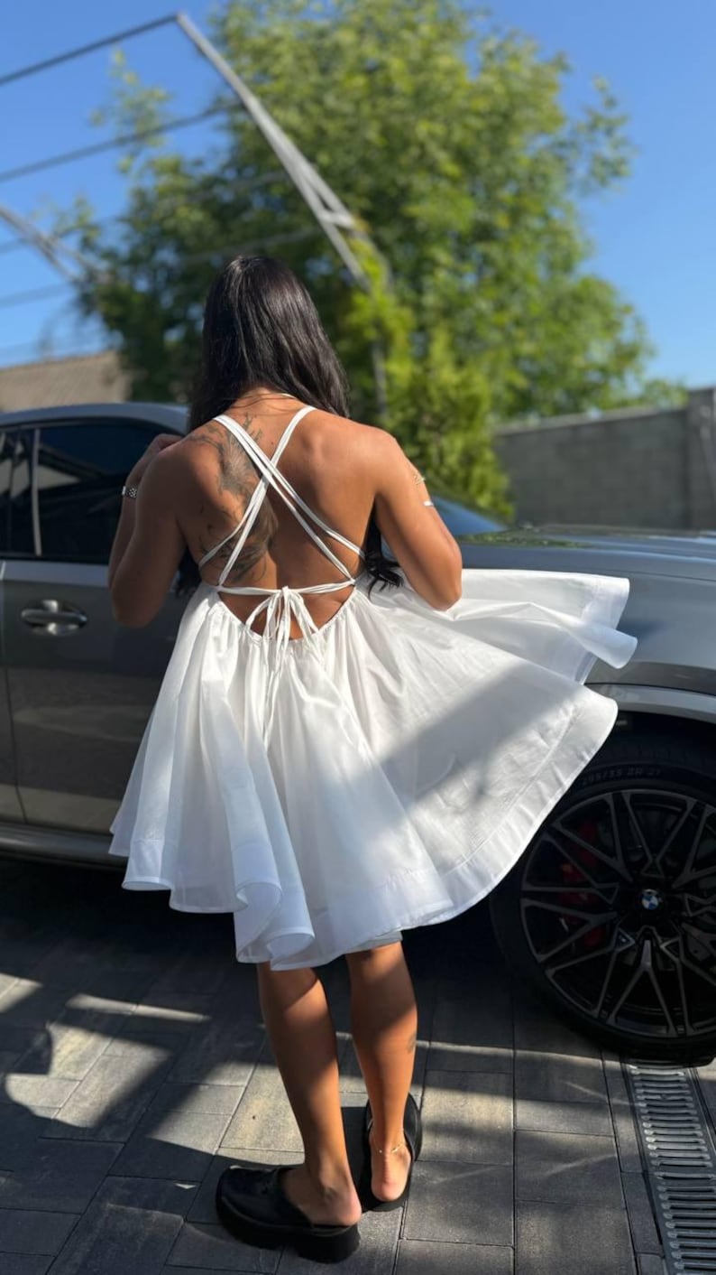 Puede incluir: Vestido blanco de algod&oacute;n con espalda cruzada y falda acampanada. El vestido llega a la rodilla y tiene un cierre de lazo en la espalda. La persona lleva sandalias de plataforma negras.