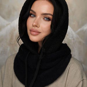 Könnte beinhalten: Schwarzer Kapuzenschal mit Kordelzug. Der Schal besteht aus einem weichen, strukturierten Material und wird über einem beigen Hemd getragen. Die Kapuze umrahmt das Gesicht und sorgt für einen warmen und stilvollen Look.