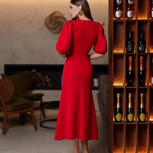 May include: A bright red midi dress with short, puffed sleeves and a fitted bodice. The dress has a flared skirt and is worn with white heels. The background includes a wooden wine rack and a fireplace.