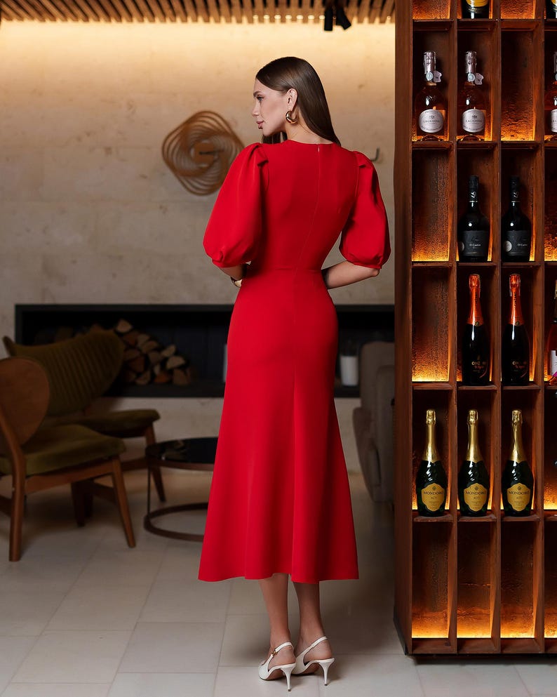 May include: A bright red midi dress with short, puffed sleeves and a fitted bodice. The dress has a flared skirt and is worn with white heels. The background includes a wooden wine rack and a fireplace.