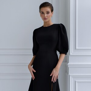 May include: A black dress featuring a round neckline and short, puffed sleeves. The dress has a fitted bodice and a long skirt with a high slit. The model is wearing gold hoop earrings and has her hair up.