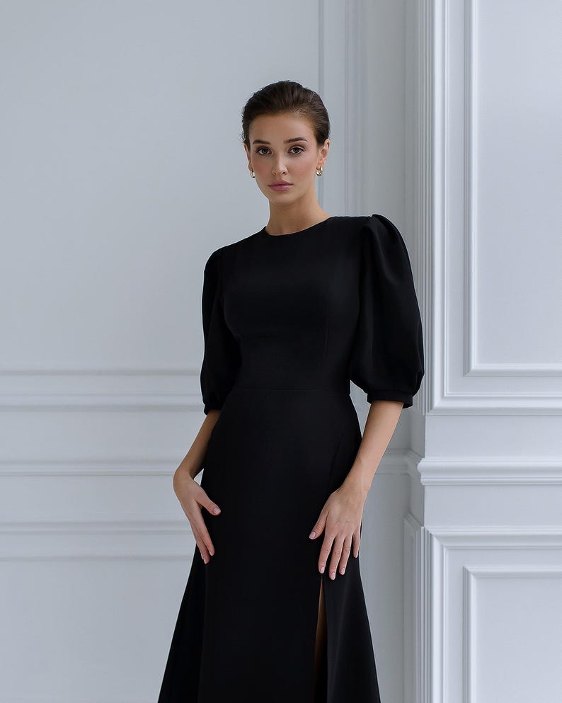 May include: A black dress featuring a round neckline and short, puffed sleeves. The dress has a fitted bodice and a long skirt with a high slit. The model is wearing gold hoop earrings and has her hair up.