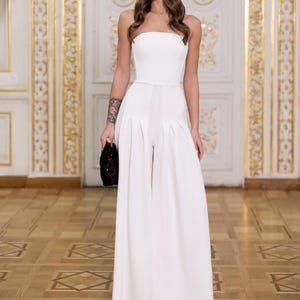 May include: A white strapless jumpsuit featuring a fitted bodice and wide-leg trousers with pleated detailing at the waist. The garment is floor-length and solid white, paired with a small black textured clutch bag held in one hand.