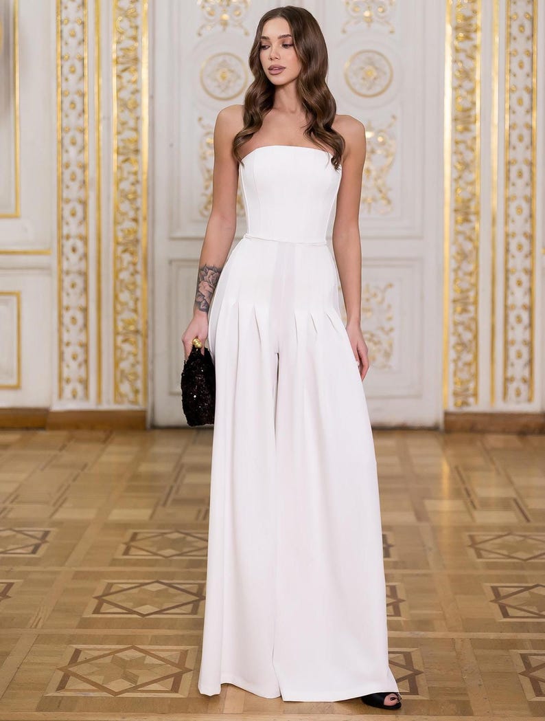 May include: A white strapless jumpsuit featuring a fitted bodice and wide-leg trousers with pleated detailing at the waist. The garment is floor-length and solid white, paired with a small black textured clutch bag held in one hand.