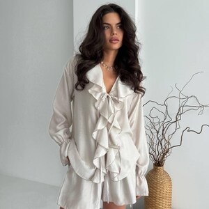 以下が含まれることがあります： フリル付きのフロントとマッチングショーツを備えた、長袖トップが特徴のライトベージュのパジャマセット。トップはVネックで、ショーツはドローストリングウエストです。セットは柔らかくシルキーな素材で作られています。