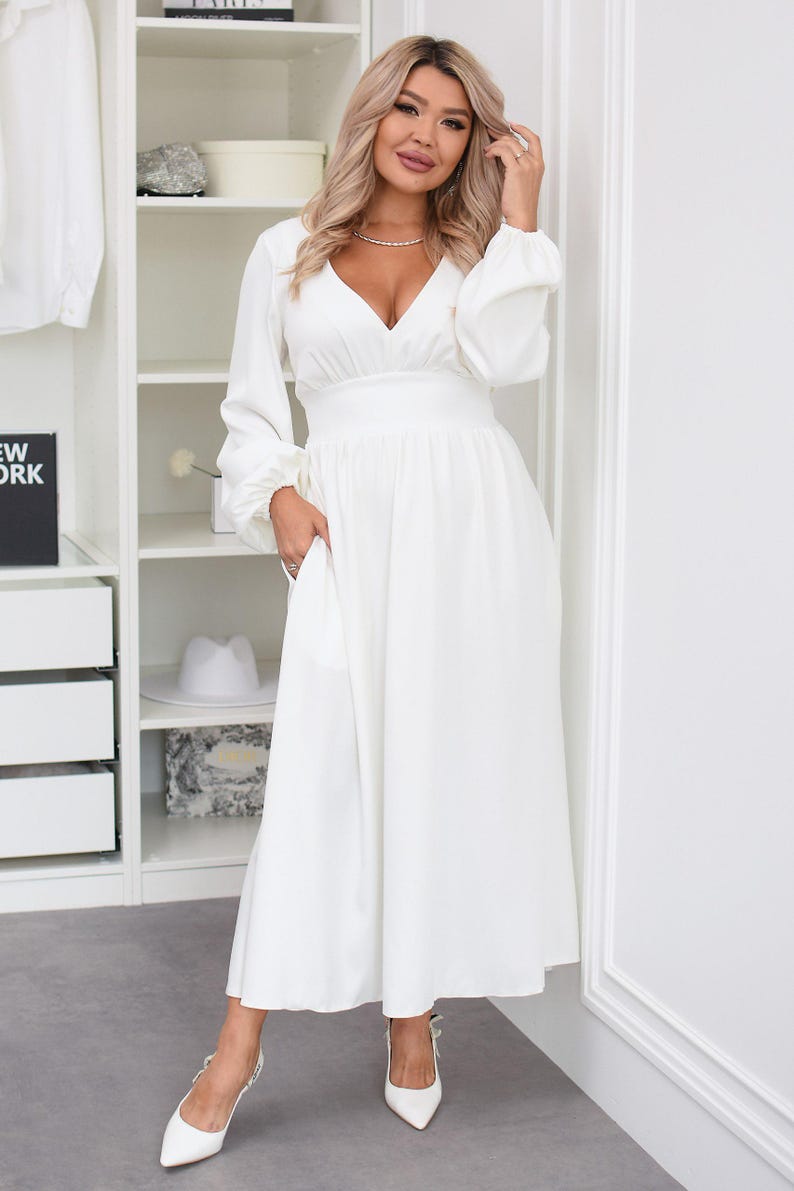 May include: A long, white dress with a V-neck and long sleeves. The dress has a fitted waist and a flowing skirt. The woman is wearing white heels. The background includes a white closet with shelves and a grey floor.