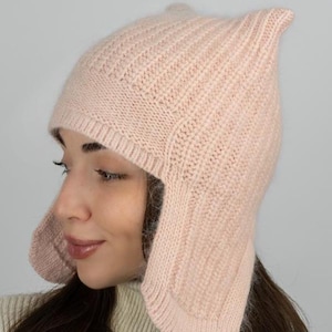 May include: A light pink knit hat featuring a ribbed texture, ear flaps, and two pointed corners at the top. The hat is constructed from a soft yarn material with a wide band around the forehead and a consistent vertical ribbing pattern throughout.