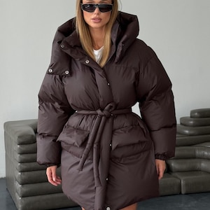 May include: A brown puffer coat with a large hood and a tie belt. The coat has a long length and a quilted design. The model is wearing sunglasses and a white shirt underneath the coat.