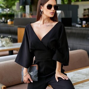 May include: A black jumpsuit featuring a deep V-neckline, wide three-quarter length sleeves, and a fitted waist. The garment is paired with a dark grey rectangular clutch bag that displays the partial white text "NE" on its front surface.