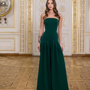 May include: A dark green strapless jumpsuit featuring a fitted bodice with vertical seam detailing and wide-leg trousers with prominent pleats at the waist. The garment has a clean, structured silhouette that extends to the floor in a solid color.