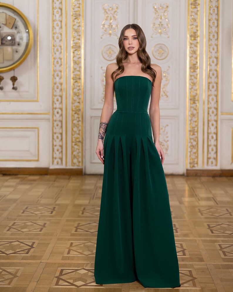 May include: A dark green strapless jumpsuit featuring a fitted bodice with vertical seam detailing and wide-leg trousers with prominent pleats at the waist. The garment has a clean, structured silhouette that extends to the floor in a solid color.