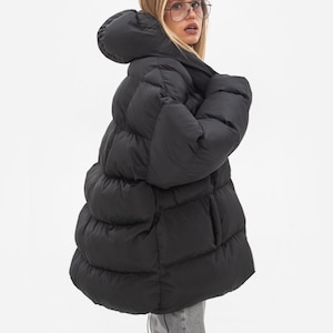 May include: An oversized black puffer jacket featuring a large hood, horizontal quilted baffles, and long sleeves. The jacket has a voluminous silhouette with a front zip fastening and is constructed from a smooth, matte-finish synthetic fabric.