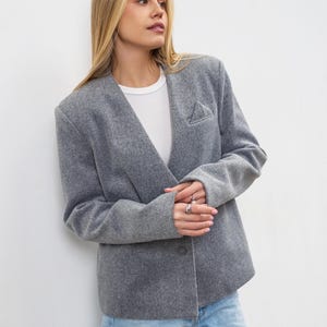 Pode incluir: Um blazer cinzento de mistura de lã com decote em V profundo, mangas compridas, fecho de um botão frontal e um pequeno detalhe de bolso triangular no peito. O blazer é usado sobre uma t-shirt branca de gola redonda e calças de ganga azuis.