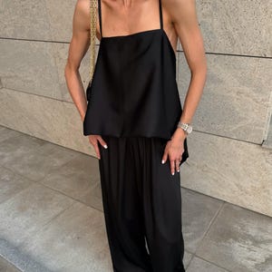 以下が含まれることがあります： 黒いサテンのキャミソールトップ、スパゲッティストラップ、お揃いのワイドレッグパンツ。ゴールドチェーンストラップバッグが肩にかかっています。この服装は、ゴールドのネックレスとシルバーの時計で装飾されています。この服装はモノクロのアンサンブルです。